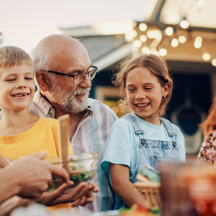 Happy Senior Grandfather Talking and Having Fun with His Grandchildren, Holding Them on Lap at a Outdoors Dinner with Food and Drinks. Adults at a Garden Party Together with Kids.