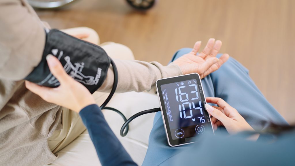 Close up woman caregiver nurse wearing blue scrubs checks blood pressure monitor and heart rate monitor with digital pressure gauge to elderly woman retirement sitting on bed in bedroom,healthcare setting medical health care plan