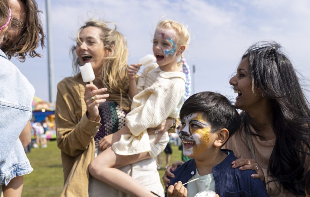 Two families visiting an Easter fair in the North East of England. They are standing together, eating ice creams and frozen lollies, enjoying the sweet treats while talking together. Some of the children have their faces painted.