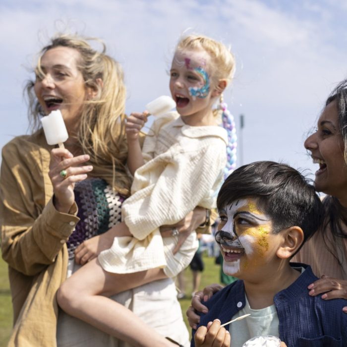 Two families visiting an Easter fair in the North East of England. They are standing together, eating ice creams and frozen lollies, enjoying the sweet treats while talking together. Some of the children have their faces painted.