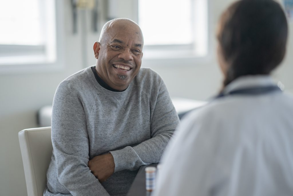 A cheerful, senior African American man sits in a clinic, sharing a warm smile with a doctor. They engage in a friendly, supportive discussion during a routine medical visit.