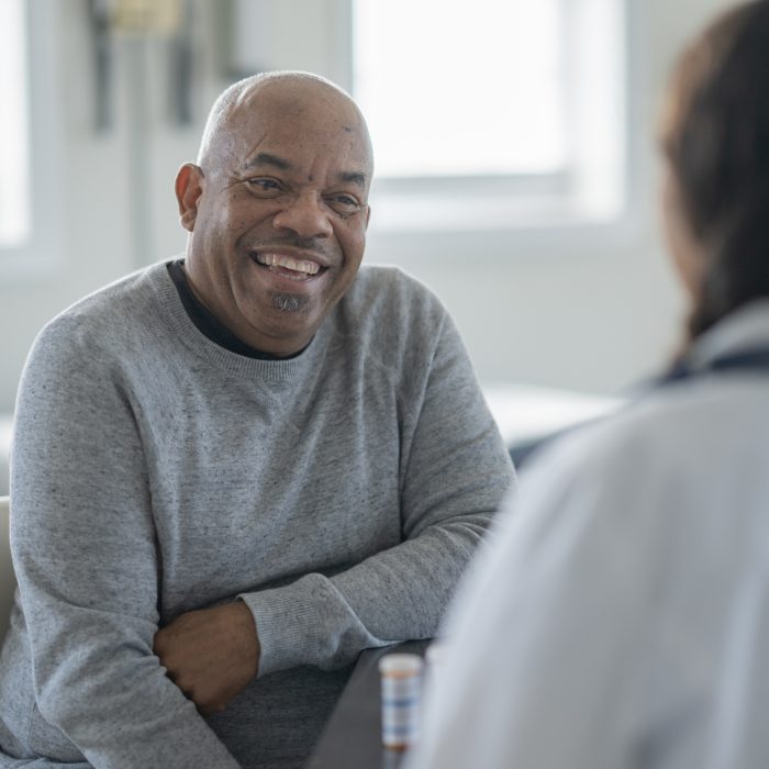 A cheerful, senior African American man sits in a clinic, sharing a warm smile with a doctor. They engage in a friendly, supportive discussion during a routine medical visit.