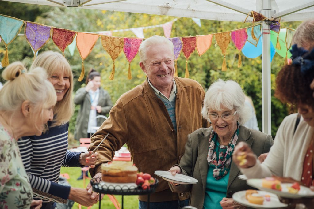 A group of seniors stand helping themselves to cakes in a garden, enjoying afternoon tea whilst chatting to eachother. There is colorful bunting hanging from above.