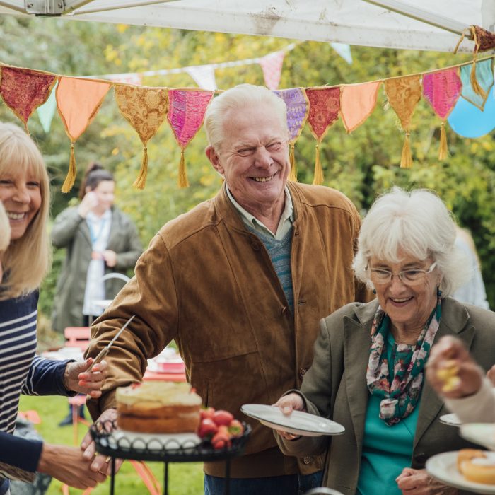 A group of seniors stand helping themselves to cakes in a garden, enjoying afternoon tea whilst chatting to eachother. There is colorful bunting hanging from above.