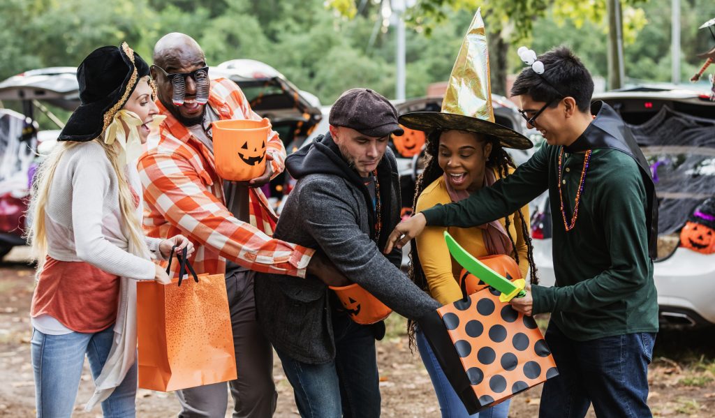 A multiracial group of five adults wearing homemade halloween costumes. They are standing in a parking lot at a trunk or treat event, reaching into one another's candy buckets.