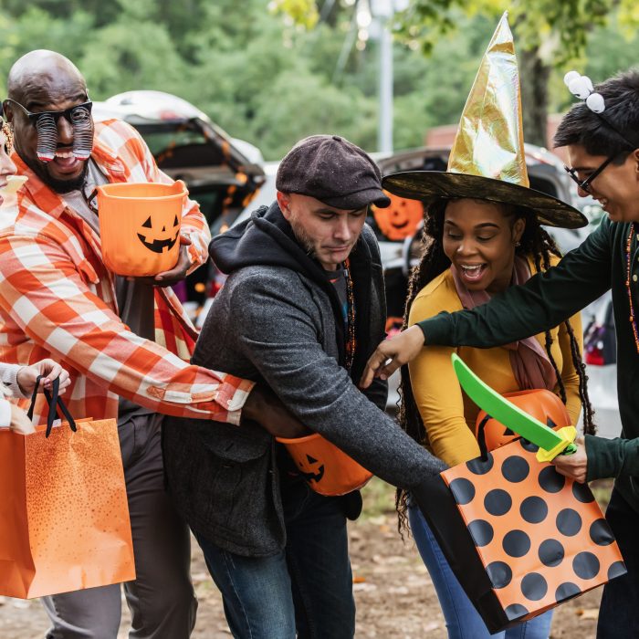 A multiracial group of five adults wearing homemade halloween costumes. They are standing in a parking lot at a trunk or treat event, reaching into one another's candy buckets.